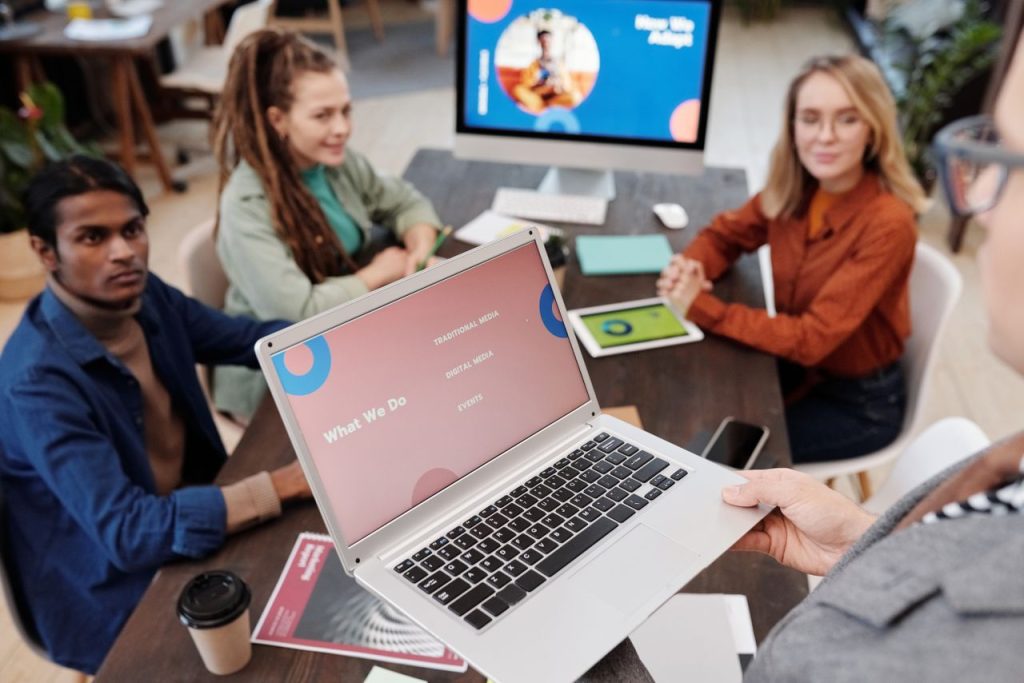 people-listening-to-the-man-holding-a-laptop-6476263 Team collaboration in an office setting with a laptop presentation and diverse colleagues.